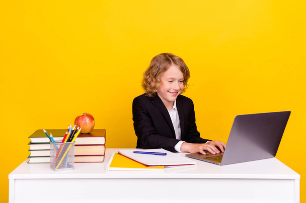 Фото занятой блондинки школьник сидеть стол делать домашнее задание типа нетбук носить униформу изолированный желтый цвет фона - Фото, изображение