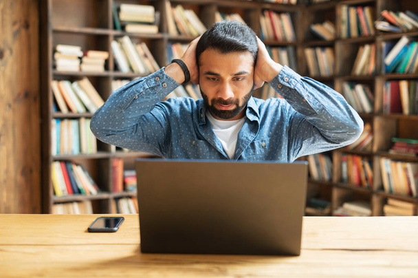 Amazed Indian male freelancer looks at laptop monitor and holding head in despair - Φωτογραφία, εικόνα