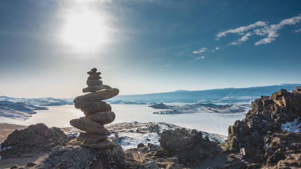 Μια πυραμίδα από πέτρες είναι χτισμένη πάνω σε ένα βράχο. Το φόντο είναι μια παγωμένη λίμνη που περιβάλλεται από μια οροσειρά. Ο ήλιος είναι στον γαλάζιο ουρανό. Μπάικαλ. - Φωτογραφία, εικόνα