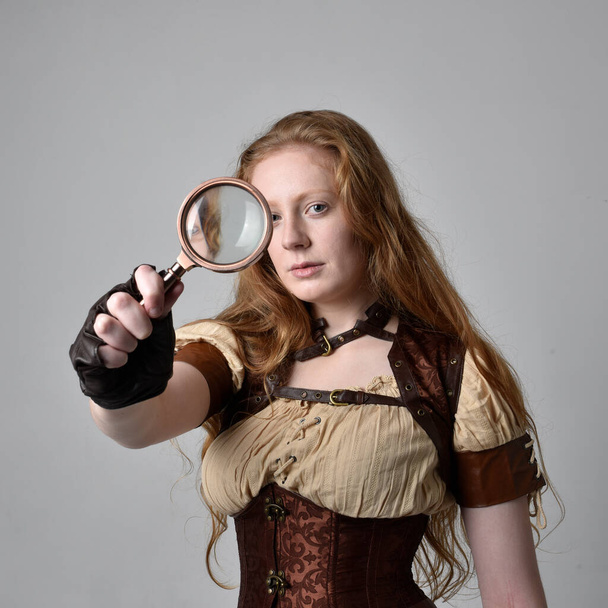 close up retrato de bela jovem com cabelo vermelho longo, vestindo steampunk inspirado traje segurando lupa isolada no fundo do estúdio. - Foto, Imagem