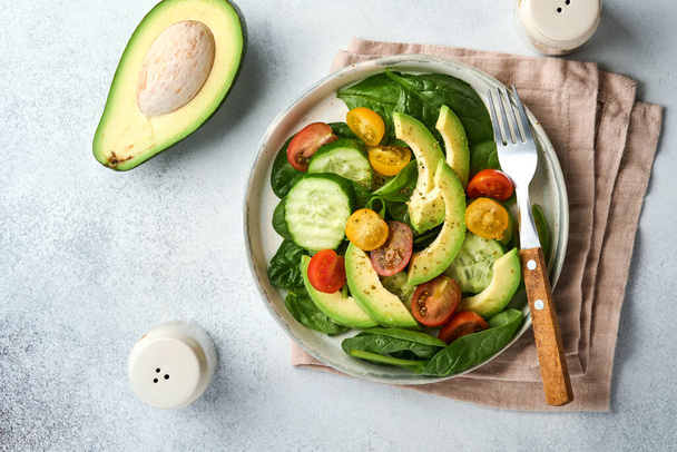 Avocado, red, yellow, black cherry tomato, spinach and cucumber fresh salad with spices pepper and olive oil in grey bowl on grey slate, stone or concrete background. Healthy food concept. Top view. - 写真・画像