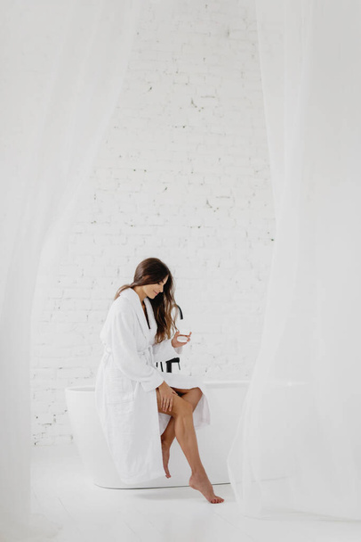 Attractive young woman sitting on bathtub and applying cream. - Фото, изображение