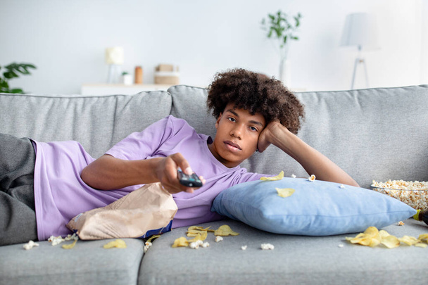 Lazy black teen guy lying on couch with scattered snacks, holding remote control, watching TV at home - Фото, зображення
