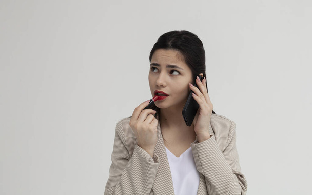 beautiful brunette in a suit paints her lips and emotionally speaks on the phone - Fotoğraf, Görsel