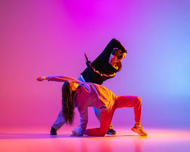 Δύο νέοι άνθρωποι, άντρας και κορίτσι με casual ρούχα χορεύουν σύγχρονο χορό, hip-hop πάνω από ροζ φόντο σε νέον φως. - Φωτογραφία, εικόνα