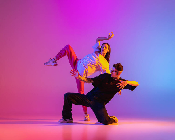 Δύο νέοι άνθρωποι, άντρας και κορίτσι με casual ρούχα χορεύουν σύγχρονο χορό, hip-hop πάνω από ροζ φόντο σε νέον φως. - Φωτογραφία, εικόνα