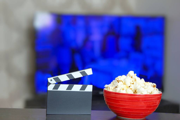 Schwarzes Klappbrett und eine Schüssel Popcorn, im Hintergrund verschwommenes Fernsehen. Hintergrundbild der Unterhaltungsindustrie. Filmzeit. Konzept des Fernsehens. - Foto, Bild