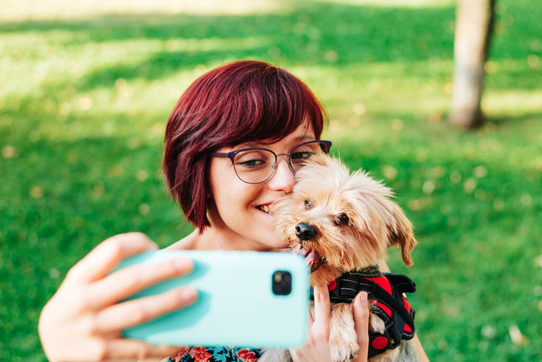 Henkilö, nuori nainen, joka ottaa valokuvan tai selfien koiransa hymyillen ja onnellisena matkapuhelimensa kanssa ulkona. - Valokuva, kuva