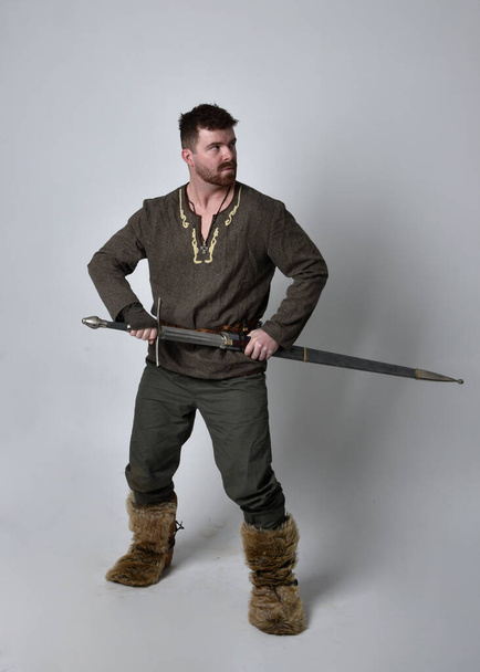 Портрет молодого красавца в средневековом костюме кельтского авантюриста. Стоячая поза, держащая меч, изолированный на фоне студии. - Фото, изображение