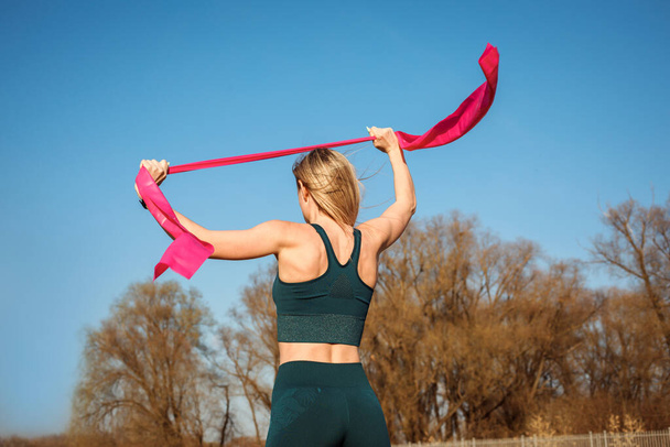 Woman is stretching her arms with elastic band, back view, exercising outdoors in spring.  - Foto, Imagem