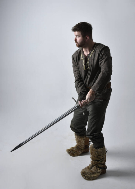 Retrato de comprimento total do jovem homem bonito vestindo traje aventureiro celta medieval. Em pé pose segurando uma espada, isolado no fundo do estúdio. - Foto, Imagem