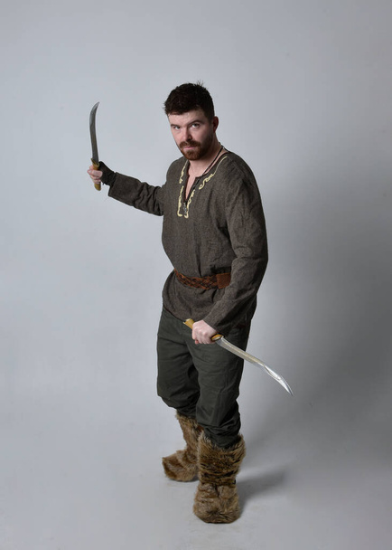 Ganztägiges Porträt eines jungen, gut aussehenden Mannes im mittelalterlichen keltischen Abenteuerkostüm. Stehende Pose mit einem Schwert, isoliert auf Studiohintergrund. - Foto, Bild
