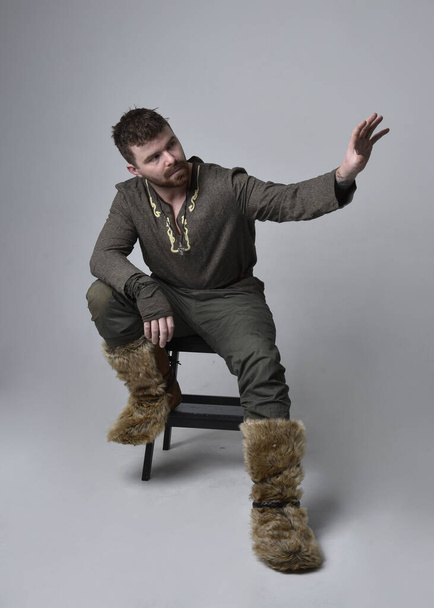 Full length  portrait of  young handsome man  wearing  medieval Celtic adventurer costume.  Sitting pose, isolated on studio background. - Photo, Image