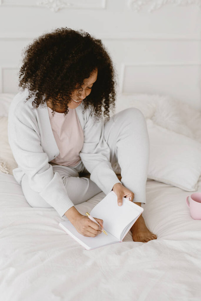 African-American woman sitting on the bed and making notes in diary. - Foto, imagen