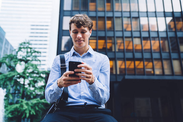 Handsome male office worker in classic blue shirt and wireless headphones messaging on phone on background of high buildings smiling - Photo, Image