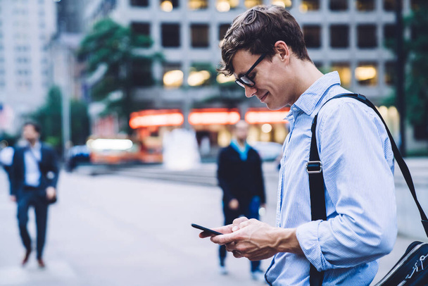 Vista lateral del trabajador de oficina en camisa y gafas con mensajería de bolsa en el teléfono de pie en el fondo de Nueva York sonriendo - Foto, imagen