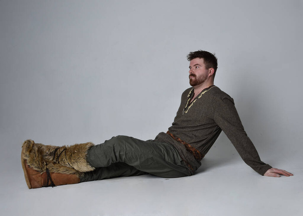 Teljes hosszúságú portré fiatal, jóképű férfiról középkori kelta kalandor jelmezben. Ülő póz, elszigetelt stúdió háttér. - Fotó, kép