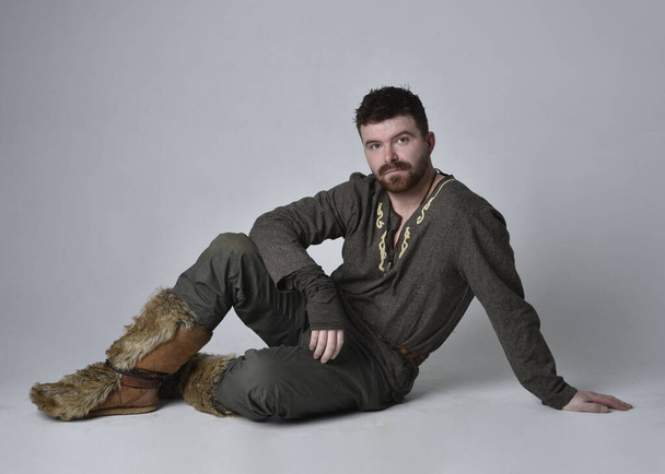 Orta Çağ Kelt maceraperest kostümü giymiş yakışıklı bir adamın tam boy portresi. Oturma pozisyonu, stüdyo arka planında izole.. - Fotoğraf, Görsel