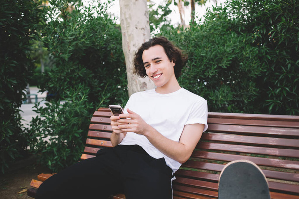 Content black haired young guy in casual wear smiling and looking at camera while sitting on bench and using mobile phone in Barcelona - Foto, Imagem