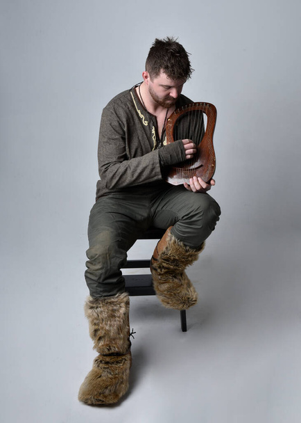 Pełen portret młodego przystojniaka w średniowiecznym celtyckim stroju poszukiwacza przygód grającego na małej harfie muzycznej, odizolowanego na tle studia. - Zdjęcie, obraz