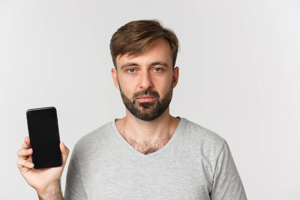 Close-up van vermoeide blanke man in grijs t-shirt, ziet er uitgeput uit en toont smartphone scherm, staande over witte achtergrond - Foto, afbeelding
