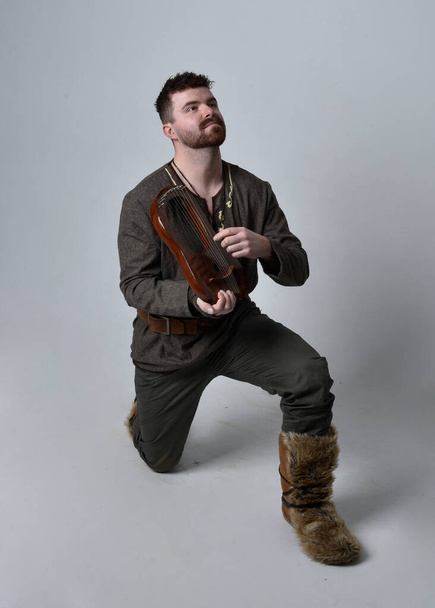 Retrato de comprimento total do jovem homem bonito vestindo traje aventureiro celta medieval tocando uma pequena harpa musical, isolado no fundo do estúdio. - Foto, Imagem