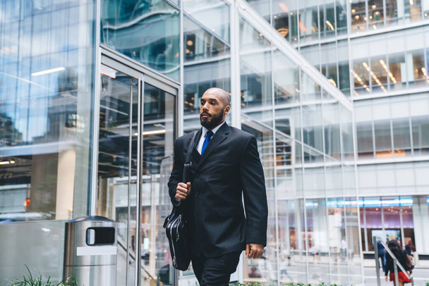 Confident bald bearded black businessman in suit holding black leather briefcase while walking in commercial building on background of glass windows  - Fotó, kép