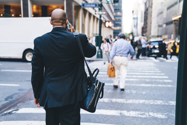 Back view unrecognizable bald African American businessman in formal wear with leather briefcase walking on street while speaking on phone - Foto, Imagen