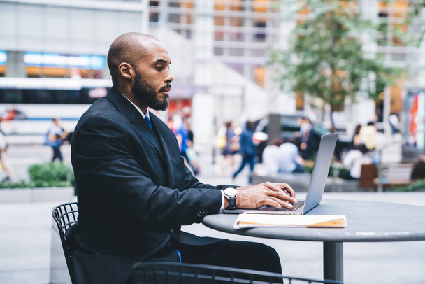 Side view of African American businessman in suit working on laptop while sitting in cafe on downtown street with folder - Φωτογραφία, εικόνα