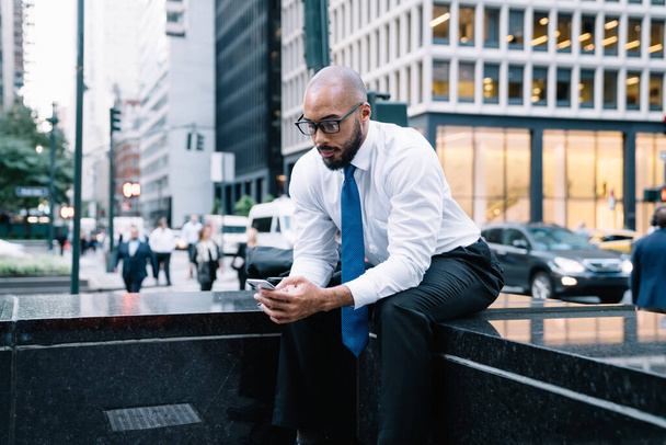 Bearded African American male in elegant clothes and eyeglasses messaging on mobile phone sitting on bench against crowded street in downtown - Foto, Imagen