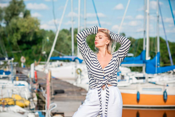 Pretty blonde woman of plus size, American or European appearance near yachts, enjoying life. Young lady with excess weight, stylishly dressed at striped jumper and white pants .Natural beauty - Zdjęcie, obraz