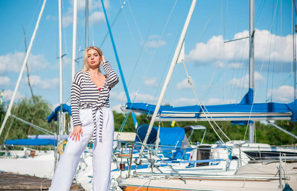Pretty blonde woman of plus size, American or European appearance near yachts, enjoying life. Young lady with excess weight, stylishly dressed at striped jumper and white pants .Natural beauty - Photo, Image
