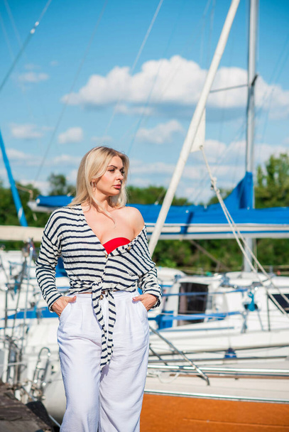 Pretty blonde woman of plus size, American or European appearance near yachts, enjoying life. Young lady with excess weight, stylishly dressed at striped jumper and white pants .Natural beauty - Фото, изображение