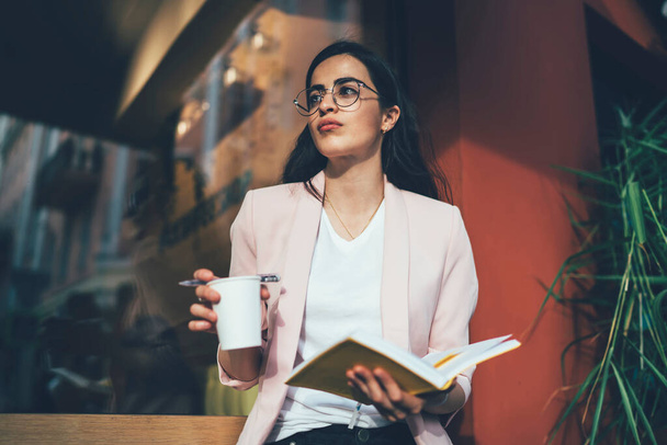 Smart casual female student with education textbook and coffee to go thinking about university text questions, contemplative woman in optical eyewear thoughtful looking away during outdoors studying - Fotoğraf, Görsel