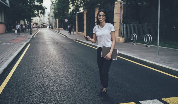Full length portrait of happy Spanish student with sketchbook and smartphone technology in hands having fun at urbanity, joyful hipster girl in spectacles posing on road and smiling at camera - Foto, imagen