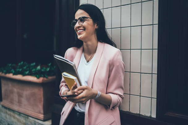 Joyful hipster student in trendy spectacles holding textbook and cellphone gadget, happy female blogger standing at urban setting and laughing during social networking on smartphone device - Photo, image