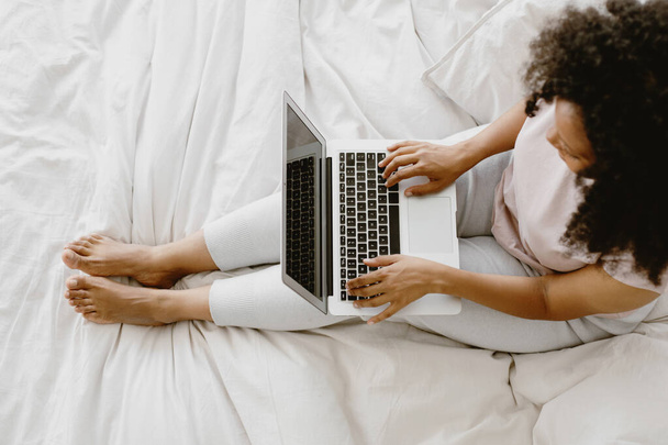 Young adorable African-American female sitting on the bed and typing on laptop. - 写真・画像