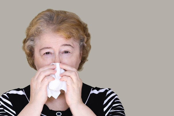 Μια ηλικιωμένη γυναίκα υποφέρει από καταρροή, κρυώνει και τρέχει η μύτη της, φυσάει η μύτη της πιέζοντας ένα λευκό μαντήλι στο πρόσωπό της. - Φωτογραφία, εικόνα