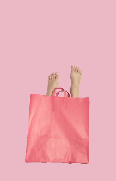 Een damesvoet komt uit een papieren roze boodschappentas op een pastelroze achtergrond. Surrealistisch shopping achtergrond concept. - Foto, afbeelding