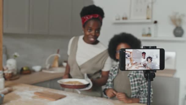 Mediana slow shot de afroamericano madre e hijo de pie por la mesa de la cocina grabación de vídeo receta de pastel de manzana casero en el teléfono inteligente - Imágenes, Vídeo