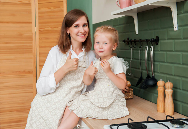 jong gelukkig mam met weinig dochter eten koekjes en show klasse in keuken thuis - Foto, afbeelding