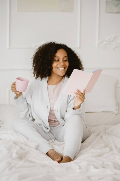 African American woman sitting on the bed reading book and drinking coffee. - Foto, Imagen