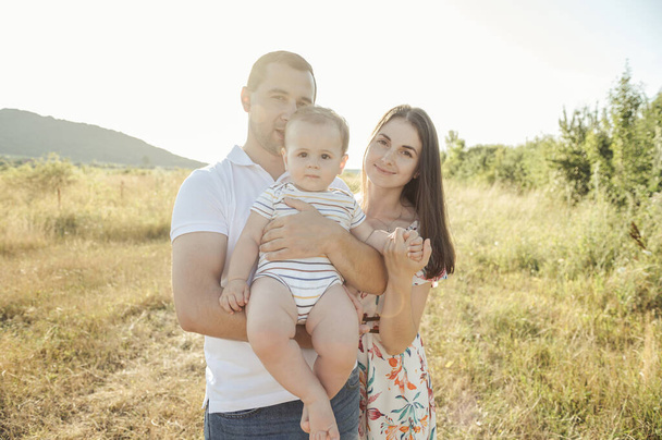Portrait of young happy family holding in hands cute toddler son enjoy sunlight at summer day on field - Foto, imagen