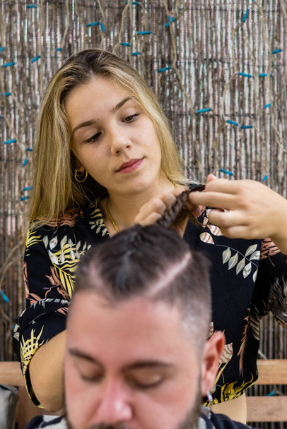 Fiatal lány hajat vág és befonja a szakállas fiatalembert - Fotó, kép