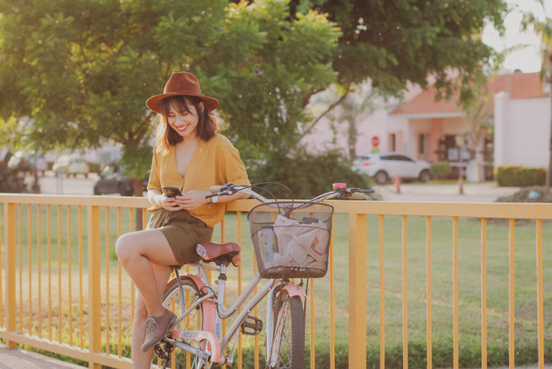Young smiling latin woman sitting on bike leaning on fence using smartphone - Φωτογραφία, εικόνα
