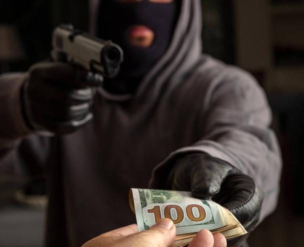 Robber in a balaclava threatens with a gun for stealing money, Armed robbery, gunman with a pistol in gloved hand, closeup view. - Фото, изображение