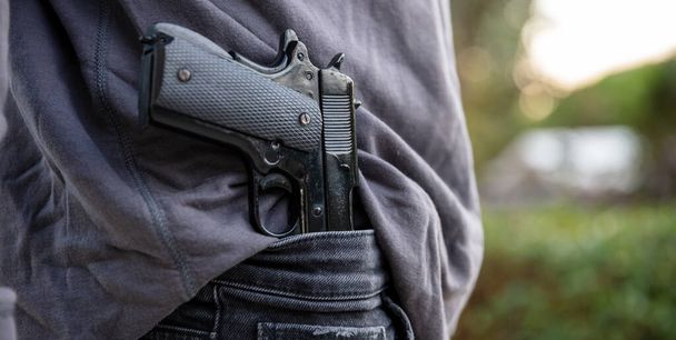 Armed man carrying a pistol in his jeans waistband, blur outdoor nature background. Threat, violence and danger concept - Photo, Image