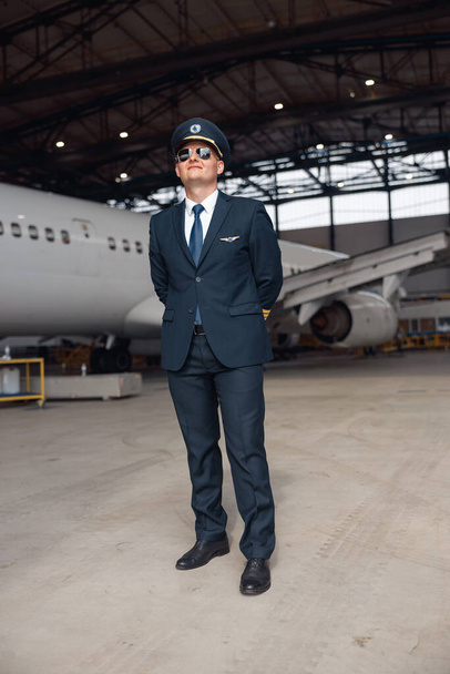 Pełna długość zdjęcia dumnego pilota w mundurze i okulary przeciwsłoneczne lotnika uśmiechającego się, stojącego przed dużym samolotem pasażerskim w hangarze na lotnisku - Zdjęcie, obraz