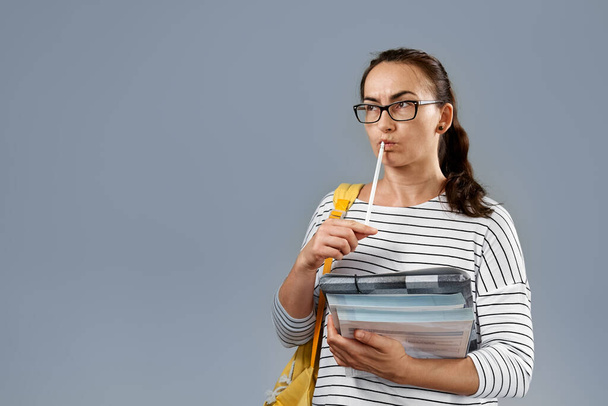 Портрет серьезной молодой женщины в очках, в полосатой футболке, с рюкзаком и книгами и держит ручку рядом с задумчивым лицом. Студентка, стоящая на фоне серой студии - Фото, изображение