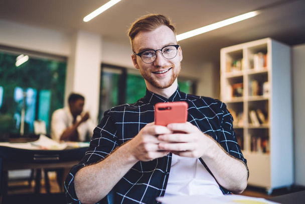 Delighted young man in casual clothes and glasses smiling and looking at camera while sitting in contemporary workspace and browsing smartphone - Fotó, kép